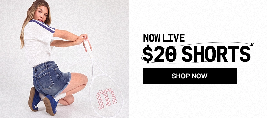 A woman kneels on a white studio background holding a tennis racket, wearing a white short-sleeve top with blue trim, a denim mini skirt, white socks, and blue sneakers. Large promotional text on the right reads 'NOW LIVE $20 SHORTS' with a black 'SHOP NOW' button.