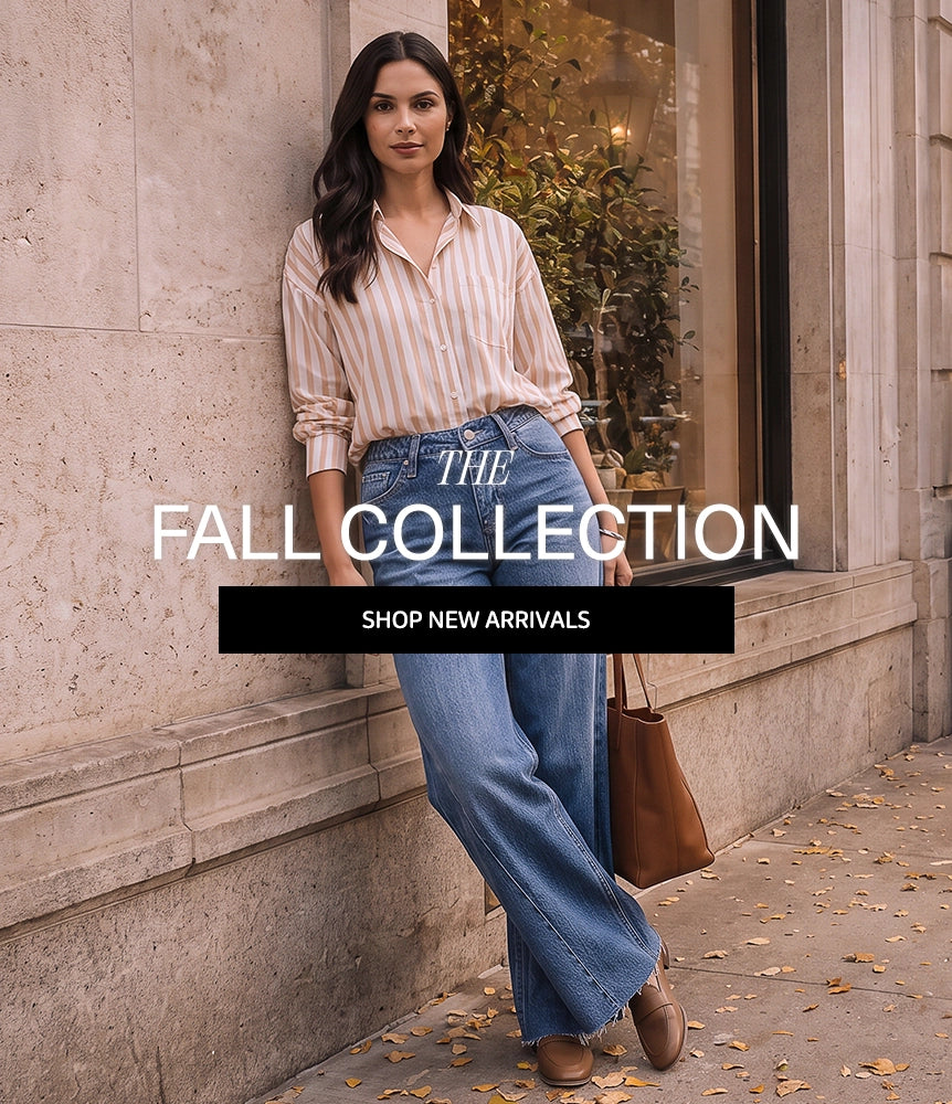 A woman stands on a city sidewalk beside a stone building window, wearing a light beige and white vertically striped button-up shirt, blue wide-leg jeans, and brown shoes, while holding a brown tote bag. Large overlaid text reads 'THE FALL COLLECTION' and 'SHOP NEW ARRIVALS'.