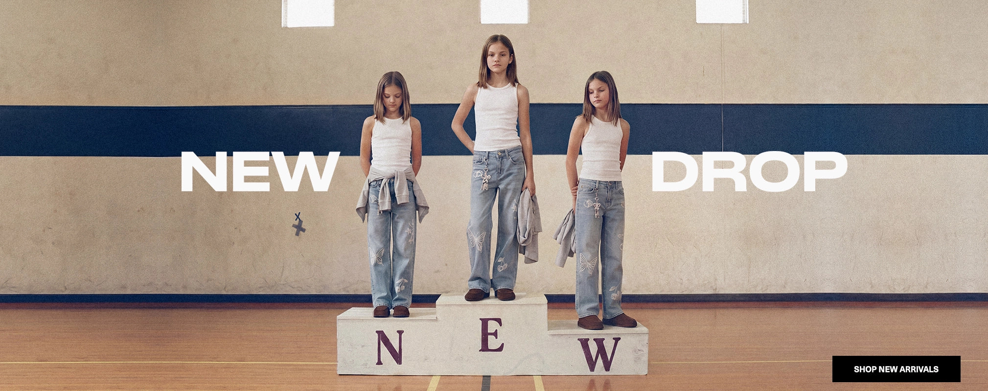 Three young girls stand on a tiered platform in an indoor gym or hall, wearing matching outfits consisting of white tank tops, light-wash wide-leg jeans, and gray sweatshirts. Large text reads 'NEW DROP' across the scene, with a small 'SHOP NEW ARRIVALS' badge in the bottom right corner.
