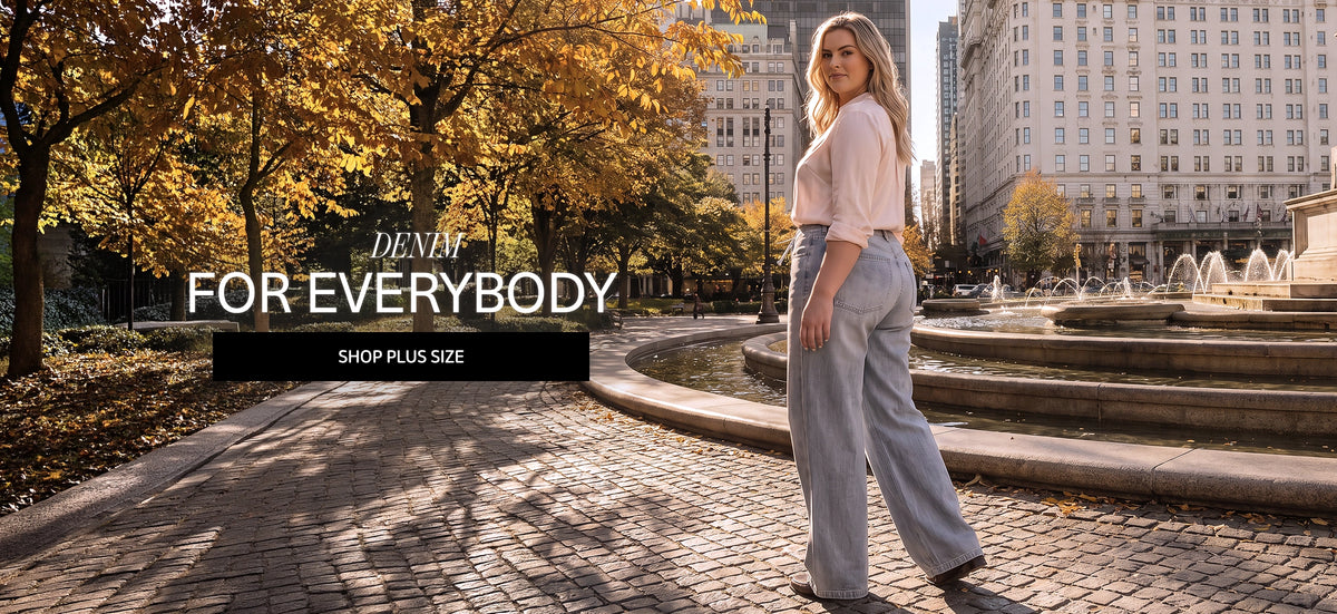 A woman stands in a city park plaza wearing a light pink short-sleeve top and light wash wide-leg jeans. Trees with yellow autumn leaves, a fountain, and buildings are in the background.