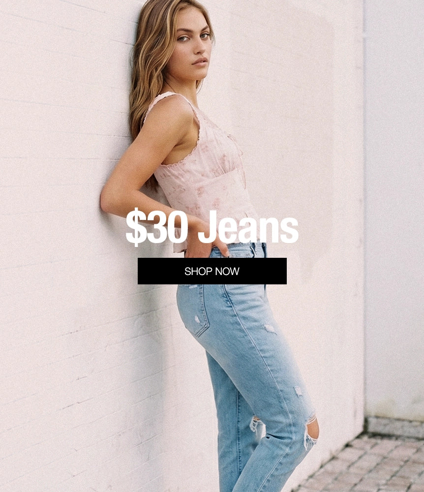 A woman leans against a white brick wall wearing light-wash distressed jeans and a pale pink sleeveless top. Large overlaid text reads '$30 Jeans' with a black 'SHOP NOW' button below.
